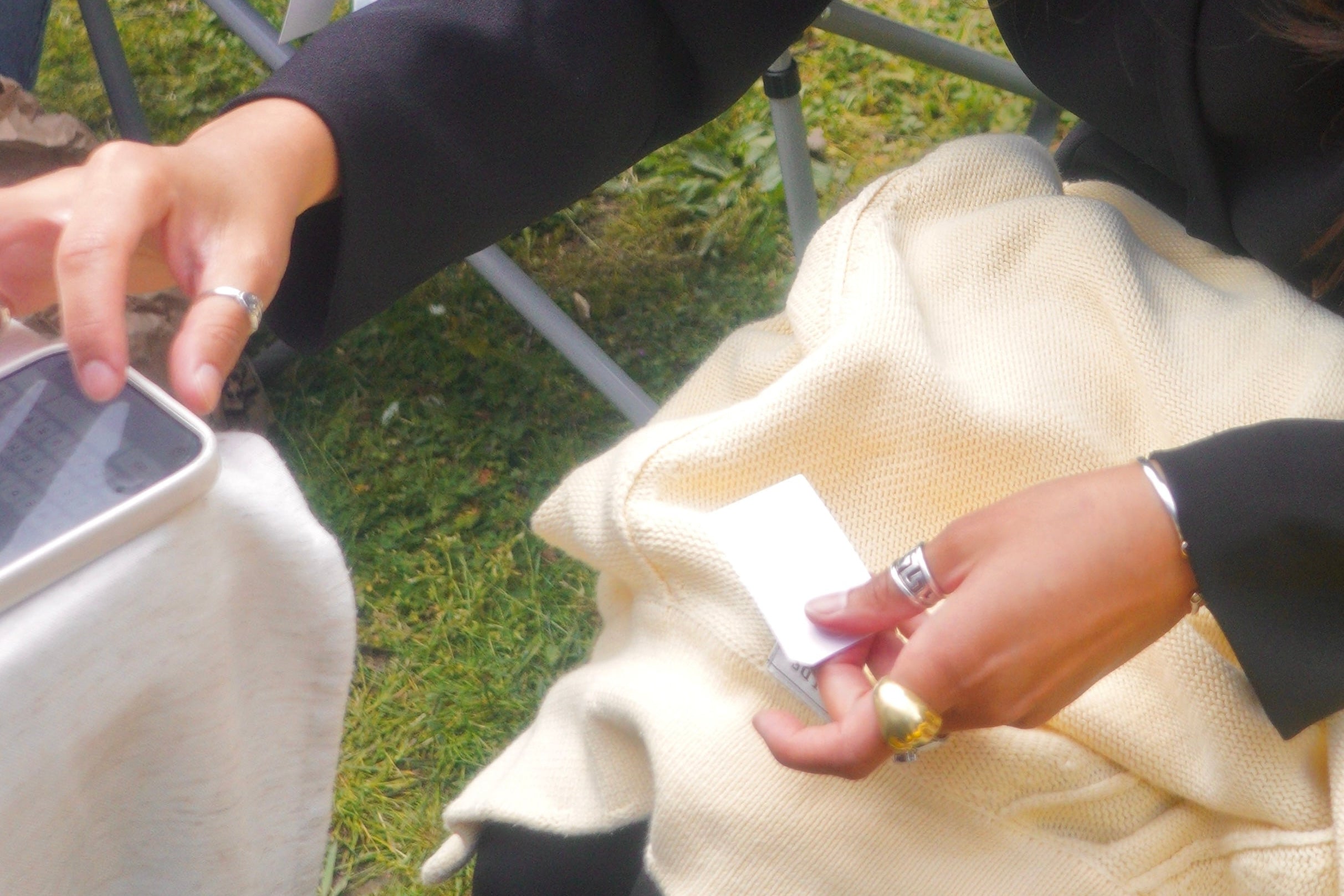 A close-up outdoor shot of a person sitting on a folding chair on grass, holding a cream-colored cardigan or sweater. The person is wearing a black top and black track pants, and has silver rings on their fingers.